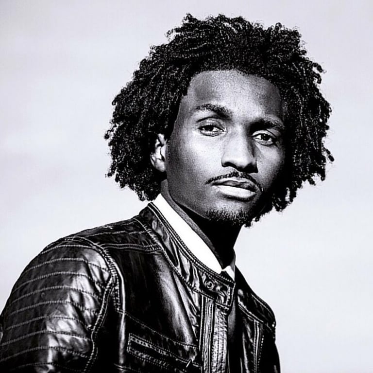 Black and white portrait of a man with curly dreadlocks and facial hair, wearing a denim jacket, facing the camera at an angle.